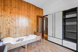 A room with a wooden wall, a bed with a white mattress, a wooden headboard, a wardrobe with open doors, and a tiled floor. - A Due Passi Dal Bosco: Hillside Apartment With Garden&bbq (Laveno Mombello)