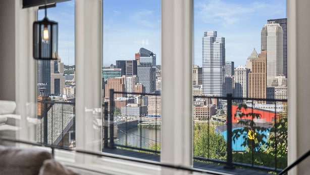 A cityscape through large windows with a view of tall buildings and a river. - Modern 3BR with Breathtaking Skyline Views (Pittsburgh)