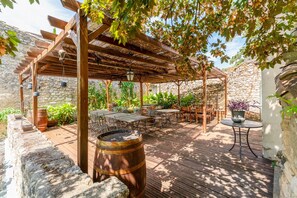 Une terrasse extérieure avec des tables et des chaises en bois, une pergola agrémentée de guirlandes lumineuses et un mur en pierre en arrière-plan.