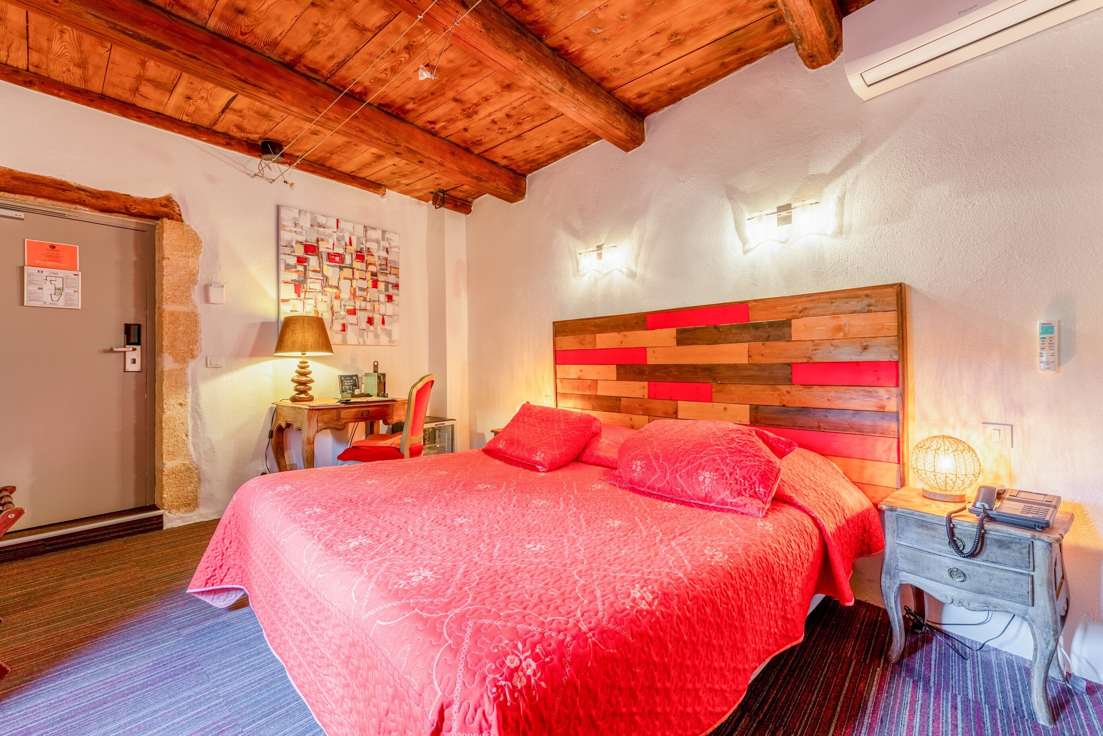 Une chambre avec un plafond en bois, un lit recouvert d’une couverture rouge, une table de chevet avec une lampe et un mur orné d’un tableau d’affichage.