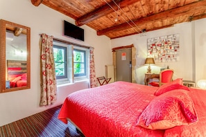 A bedroom with a wooden ceiling, a bed with red bedding, a window with curtains, a desk with a lamp, and a wall with a bulletin board.