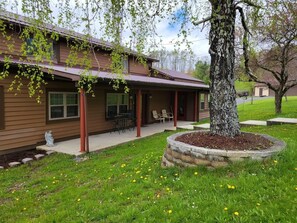 A two-story house with a covered porch, a stone planter with a tree, and a lawn with dandelions. - Hot Tub + Fishing Pond + King Suite Retreat (8-10) (Thomas)