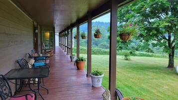 Un área de estar al aire libre techada, con mesas y sillas, plantas en macetas y vistas a un paisaje cubierto de césped.