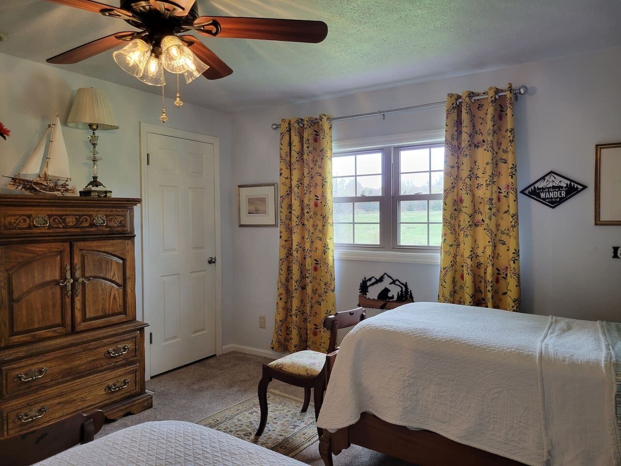 A bedroom with two beds, a wooden dresser, a chair, a ceiling fan, and a window with curtains.