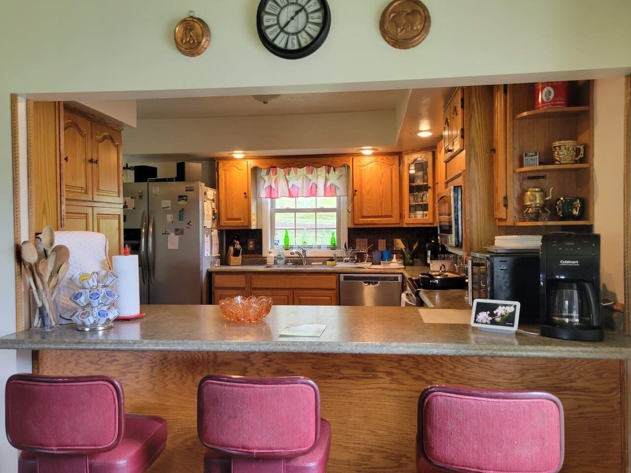 A kitchen with wooden cabinets, a refrigerator, a coffee maker, and a window with curtains.