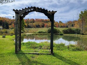 A gazebo with a lattice top, situated in a grassy area with a pond and trees in the background.