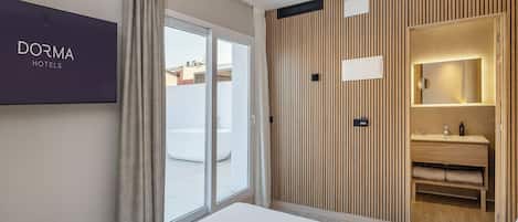 A modern hotel room with a large bed, a sliding glass door to a balcony, a wooden panel wall, and a mounted television displaying 'DORMA HOTELS'.