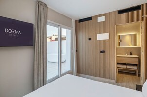 Una habitación de hotel moderna con una cama grande, una puerta corrediza de vidrio a un balcón, un panel de pared de madera y un televisor con el letrero 'DORMA HOTELS'.