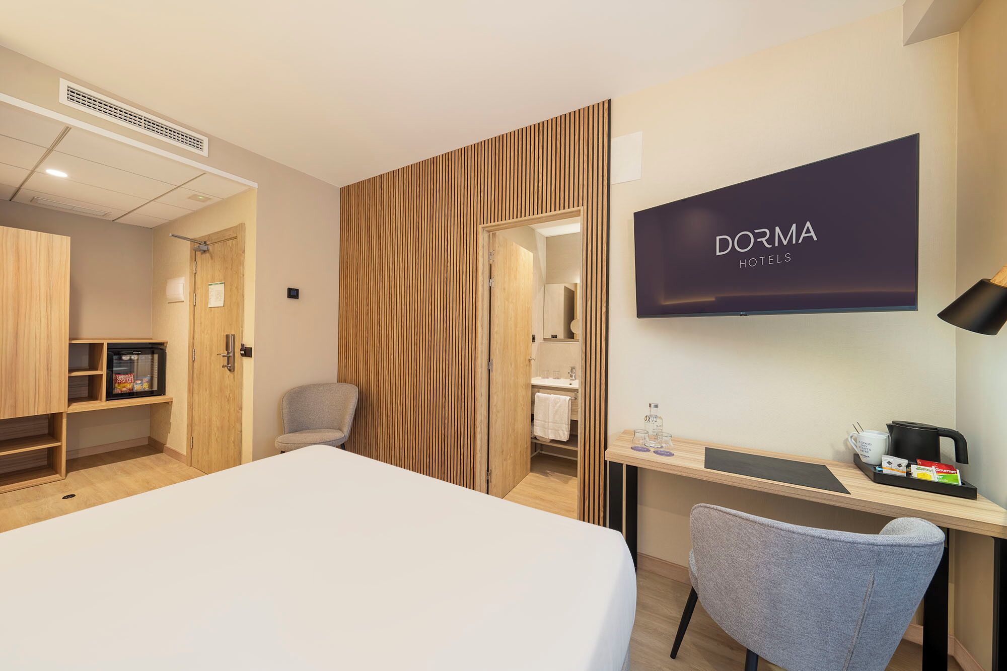 A hotel room with a bed, a desk with a chair, a TV, and a minibar.