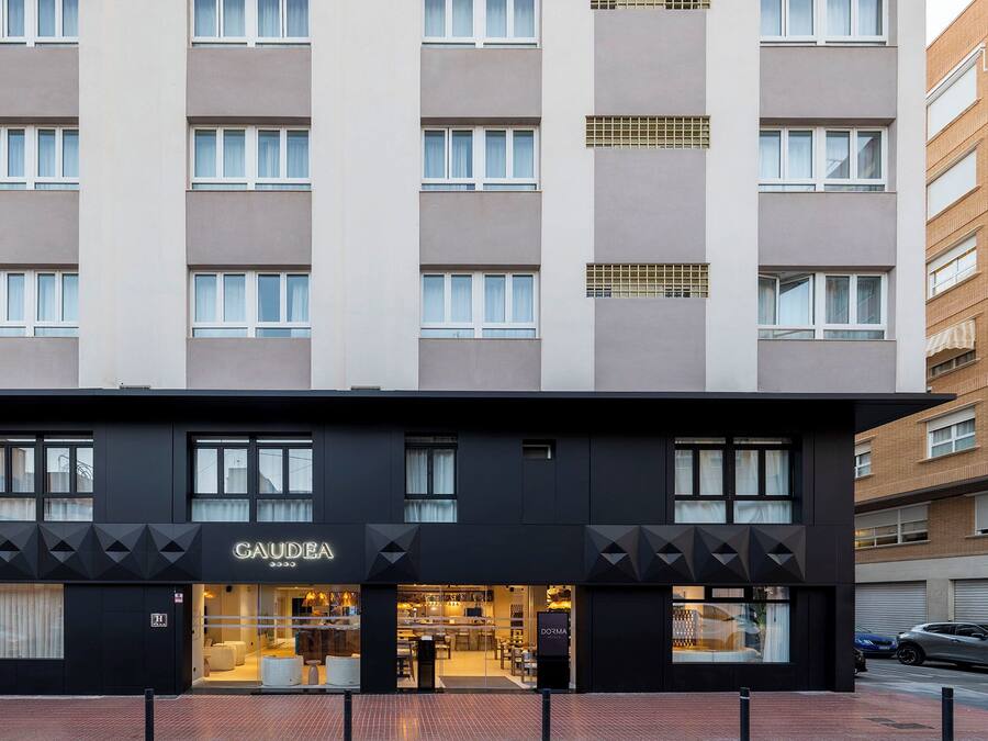 A modern hotel with a black facade, multiple windows, and a visible store named 'GAUDEA' on the ground floor.