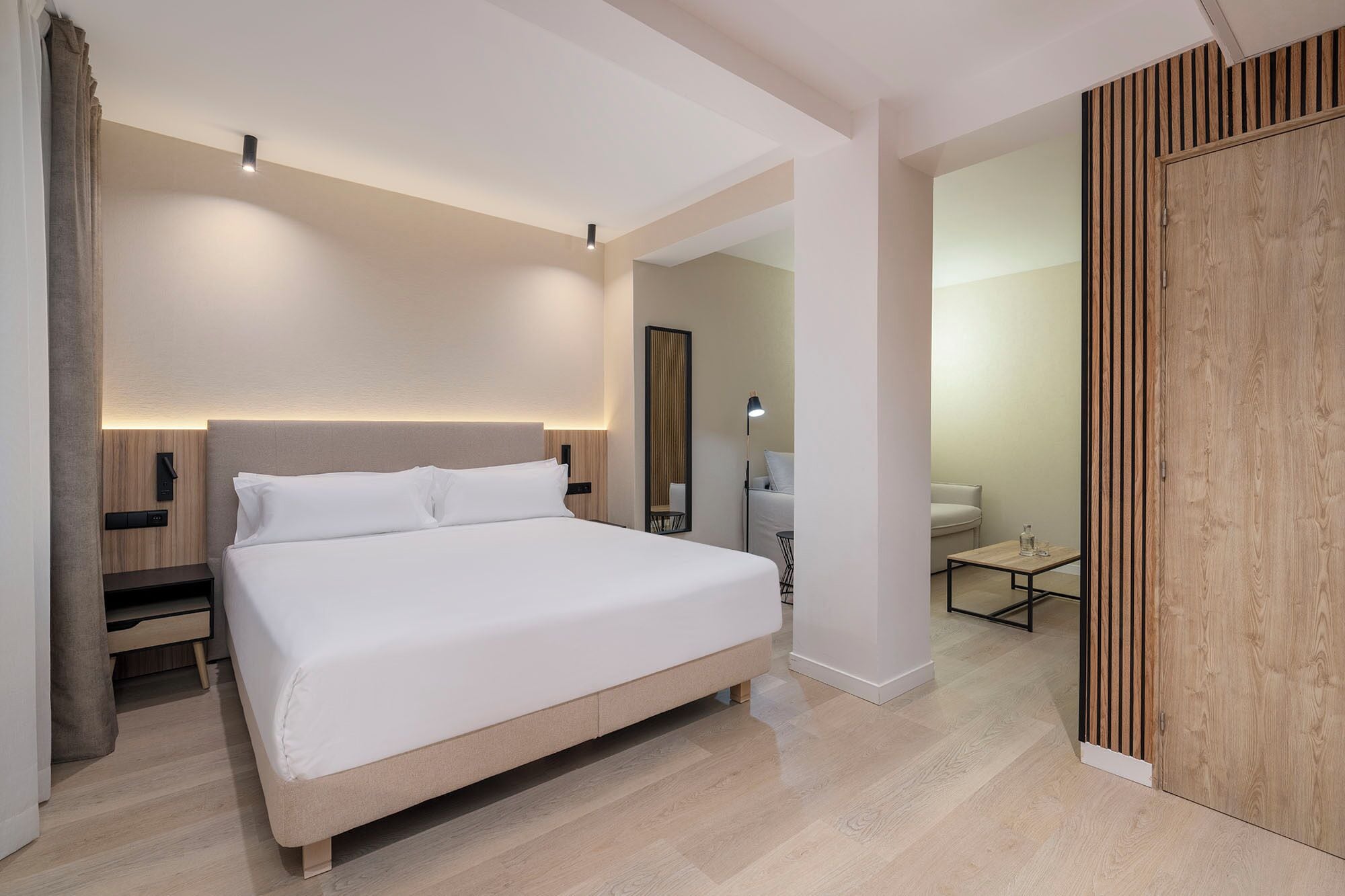 Een moderne hotelkamer met een groot bed, bedieningsknoppen langs het bed, een nachtkastje, een spiegel, een klein tafeltje met een lamp en een houten scheidingswand.