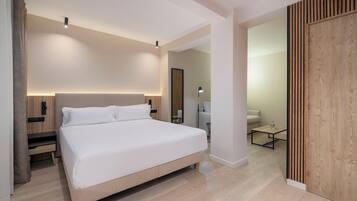 A modern hotel room with a large bed, bedside controls, a nightstand, a mirror, a small table with a lamp, and a wooden partition.