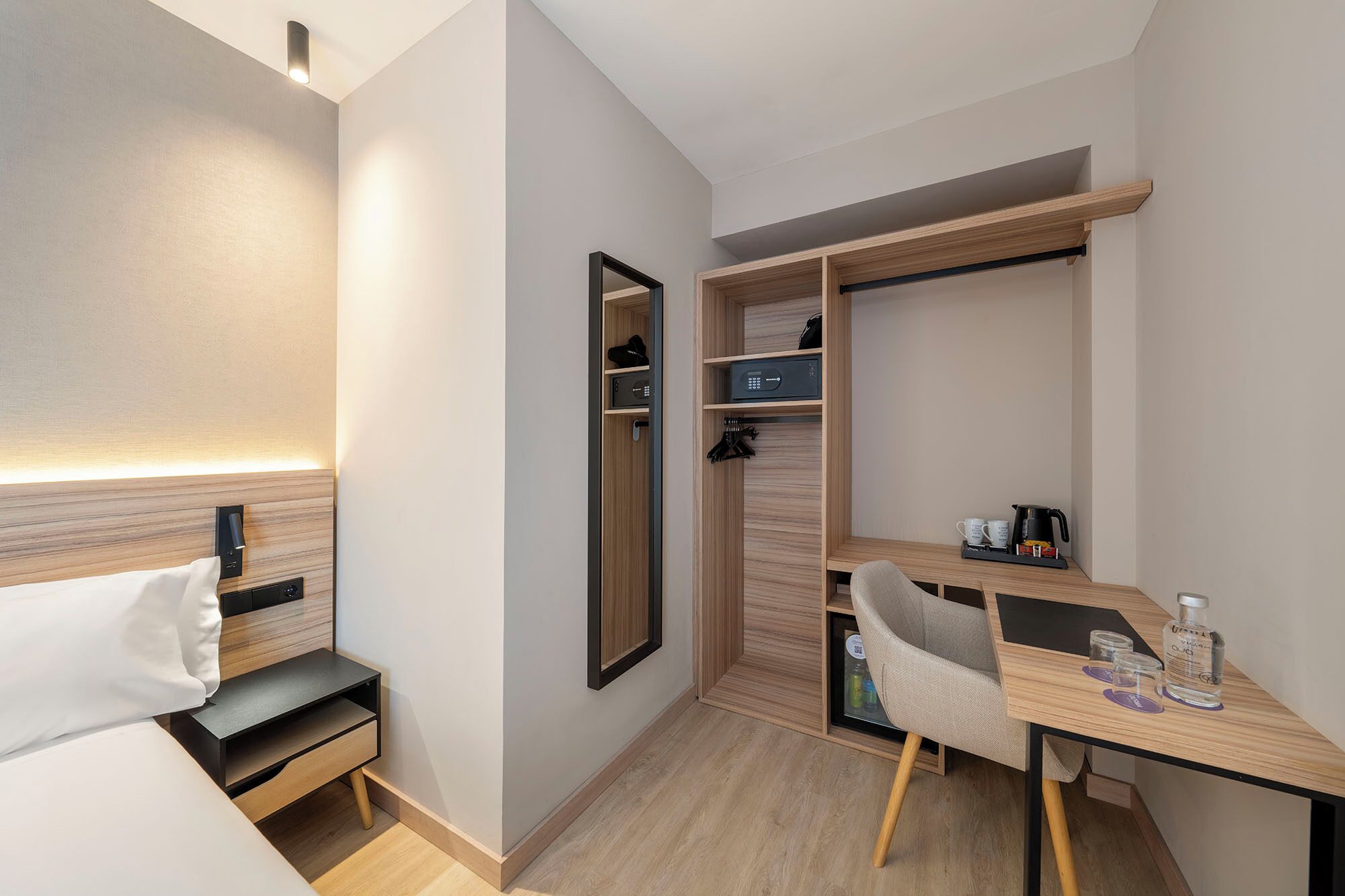 A compact hotel room with a bed, a desk, a chair, a wardrobe, and a mirror.