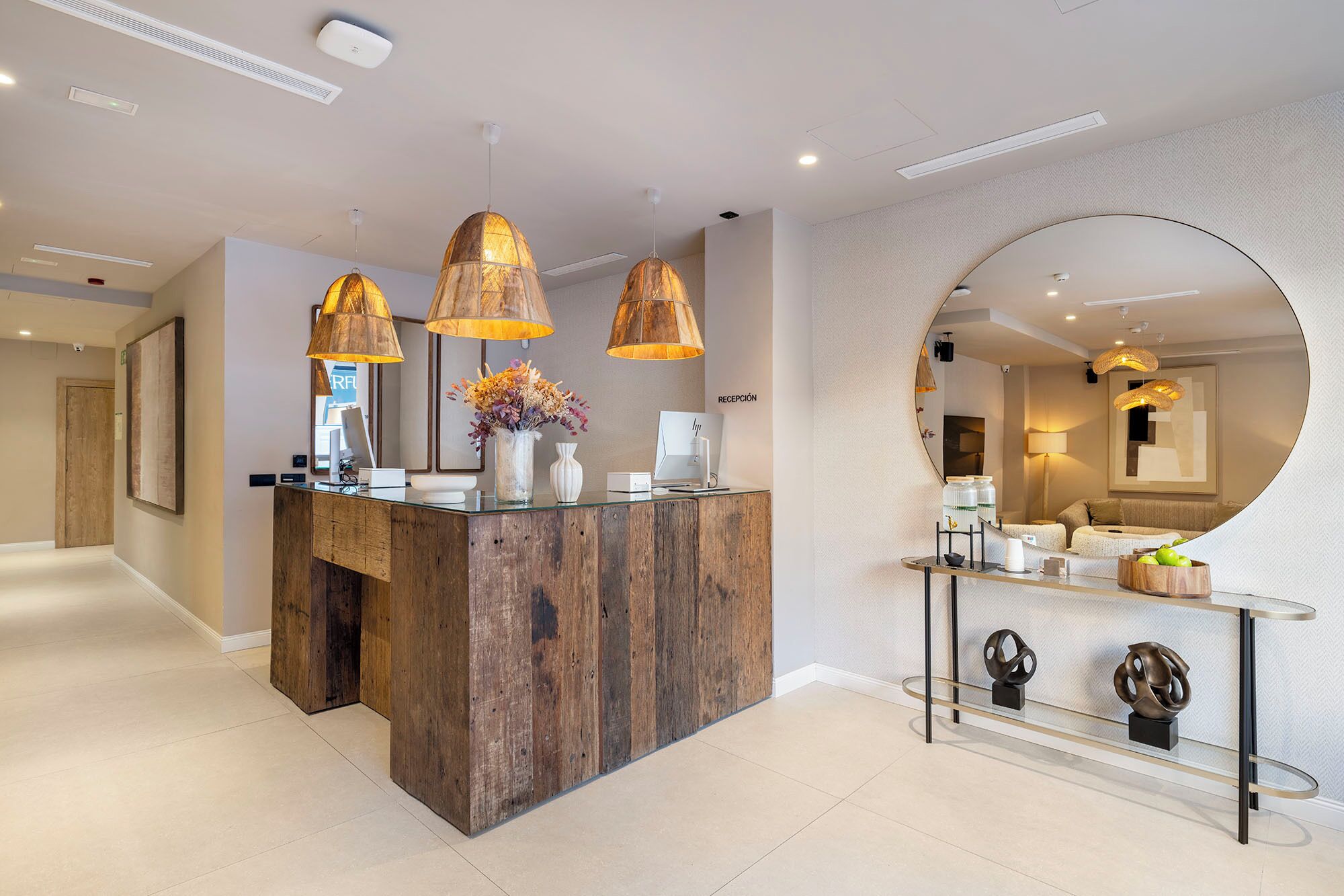 Zona de recepción con un mostrador de madera, tres lámparas colgantes, un espejo grande y una vitrina de vidrio con objetos decorativos.