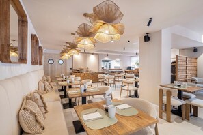 Un comedor moderno con mesas y sillas de madera, una lámpara central y una cocina al fondo.