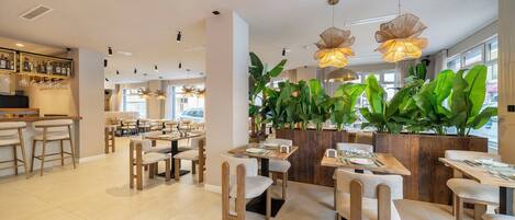A restaurant interior with multiple dining tables and chairs, a bar area with bottles and glasses, and large green plants.