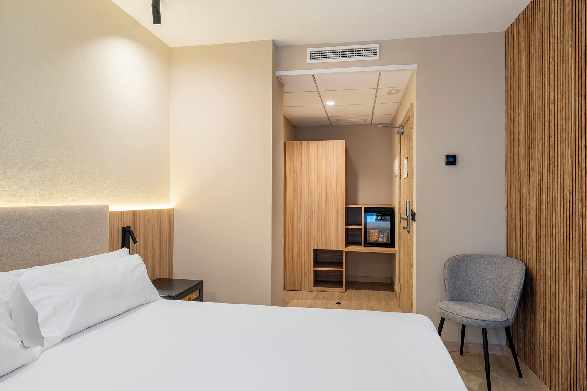 A hotel room with a bed, a chair, a wardrobe, and a small kitchenette.