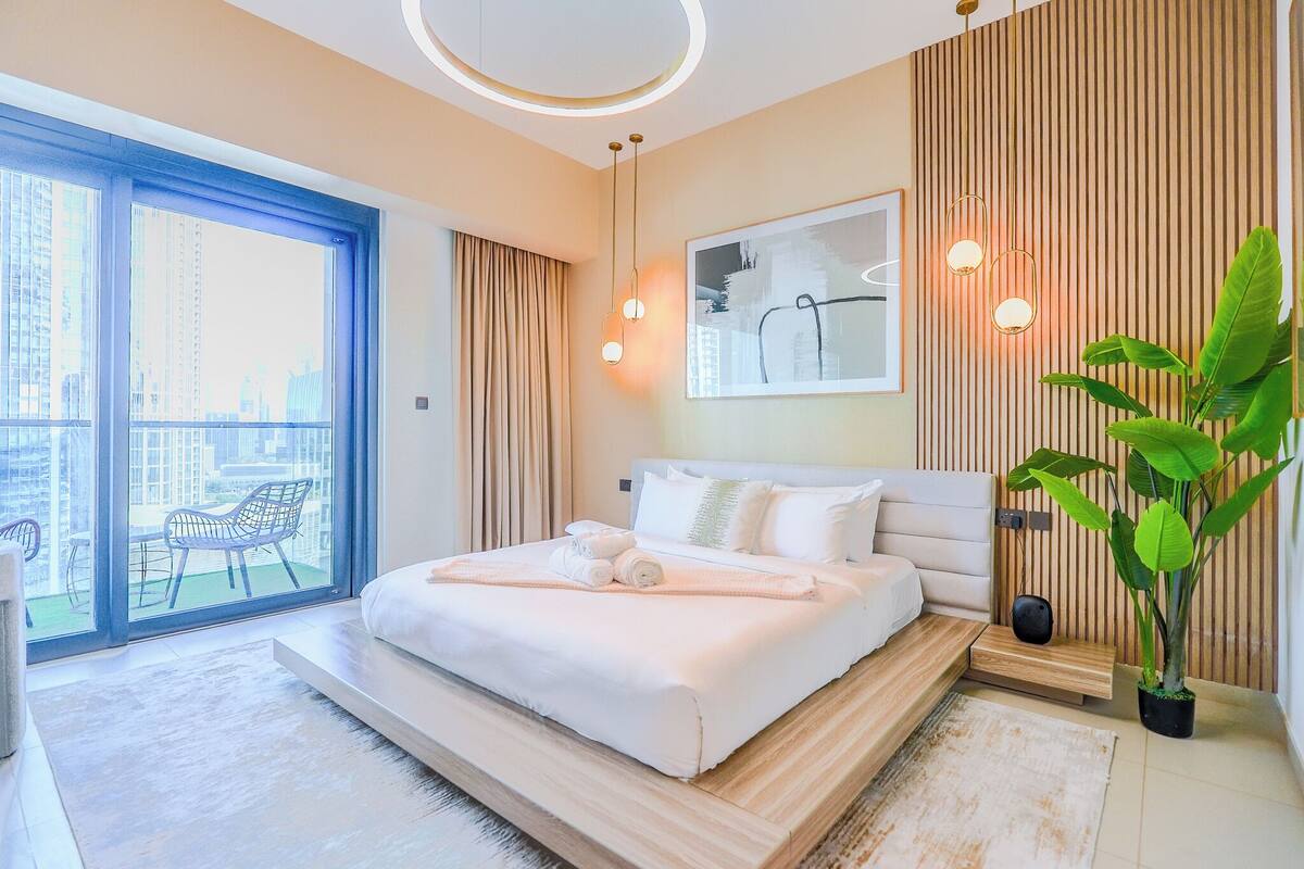 Ein modernes Schlafzimmer mit einem großen Bett, Blick auf die Stadt durch eine Schiebetür und einer Zimmerpflanze.