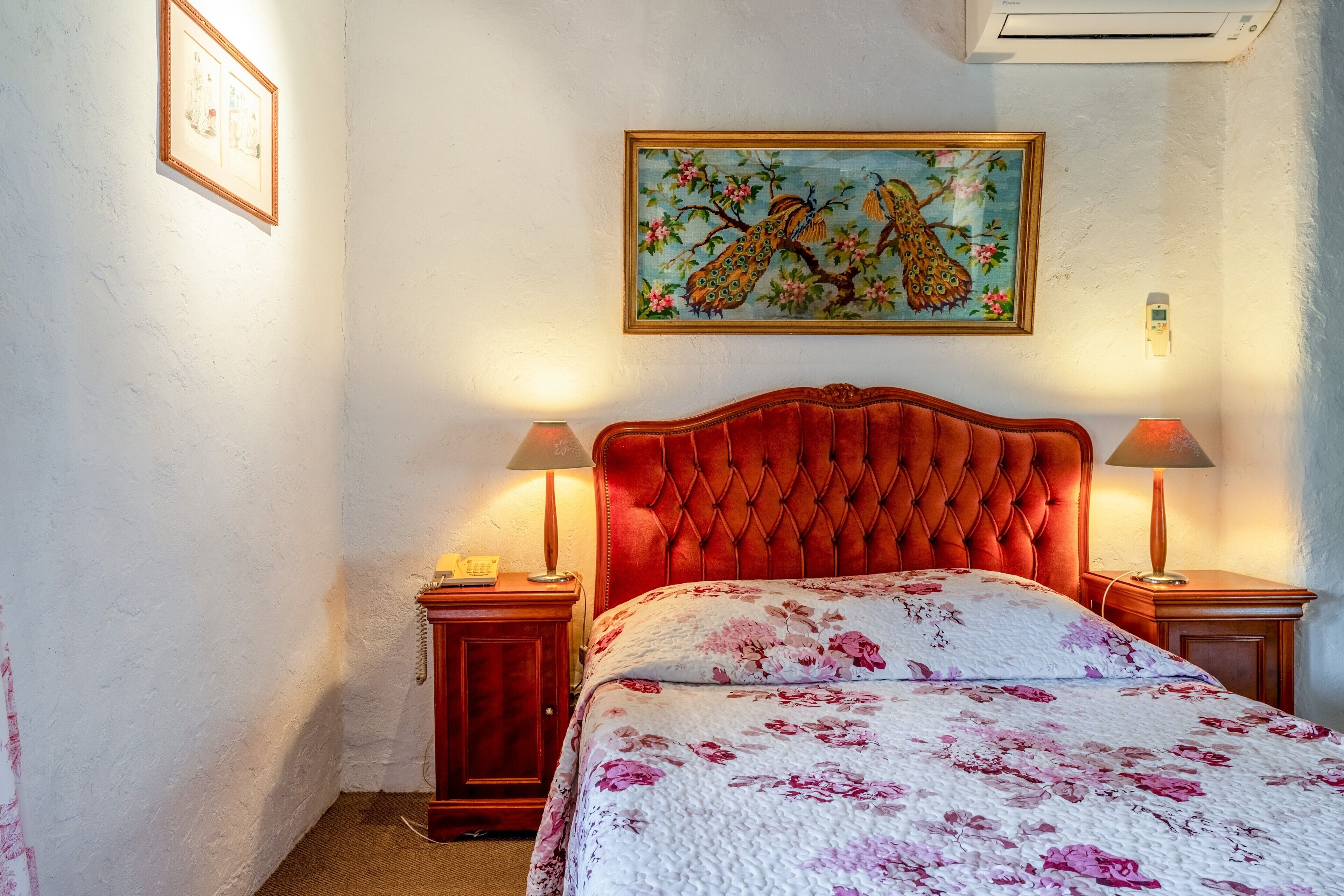 Une chambre à coucher avec une tête de lit capitonnée, une literie à motifs floraux, des lampes de chevet et un climatiseur fixé au mur.