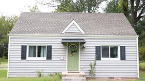 A single-story house with a green front door, two windows with shutters, and a small porch.