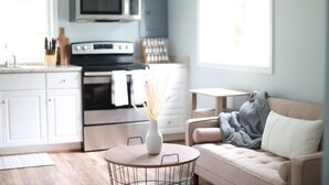 A modern kitchen with white cabinets, a microwave, and a stove. There's a dining area with a round table and chairs, and a sofa with a blanket.