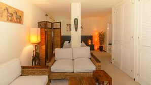 A living room with a sofa, a wooden coffee table, and a lamp. There is a bed with a headboard and a bedside table with a lamp. The room has white walls and a large window. - WELCS APARTMENT 284 PDA with terrace and view (Sant Feliu de Guixols)