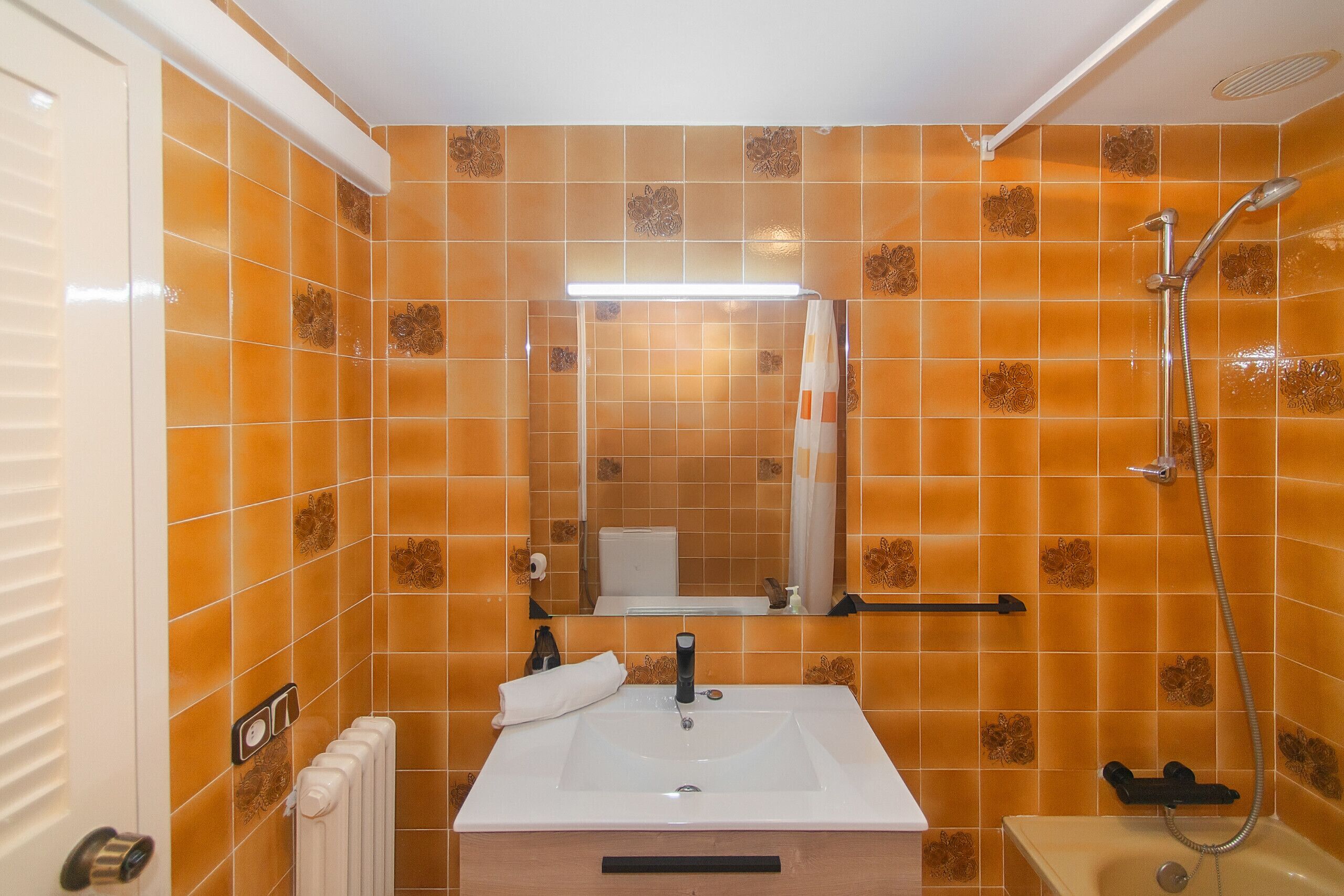 Une salle de bain avec des carreaux orange, un lavabo blanc, un miroir, une serviette et une douche.