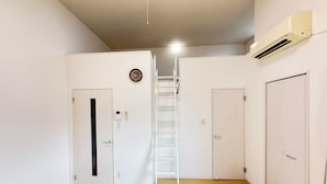 Una habitación con una puerta blanca, un aire acondicionado blanco instalado en la pared, una escalera blanca y suelo de madera.