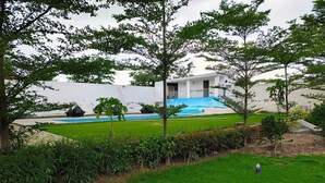 Une piscine entourée d’une pelouse, avec des arbres et un bâtiment blanc en arrière-plan.