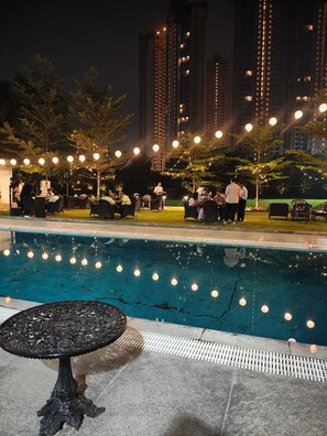 Zona junto a la piscina con luces colgantes, asientos exteriores y vista a edificios altos al fondo.
