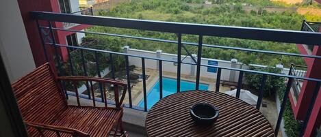 Ein Balkon mit zwei Holzstühlen und einem kleinen runden Tisch, mit Blick auf ein Schwimmbad und eine grüne Landschaft.