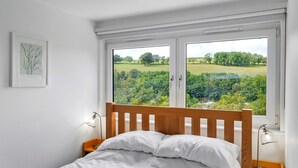 A bedroom with a wooden headboard, white bedding, and a view of a green landscape through large windows. - Three Quarter Cottage (Bodinnick)