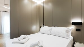 A modern hotel room with a neatly made bed, a bedside table, and a wall-mounted light fixture. - Bfor Suite Experience (Nola)