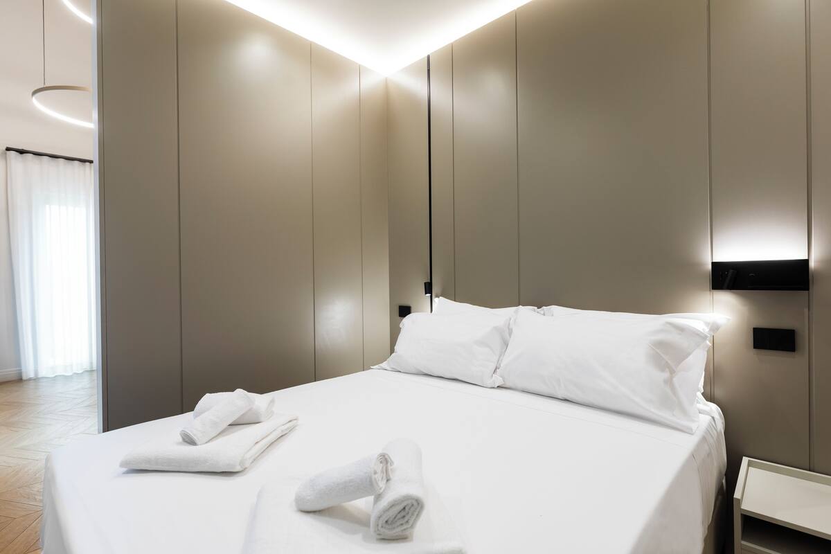 Quarto de hotel moderno com uma cama bem arrumada, mesa de cabeceira e luminária fixada na parede.