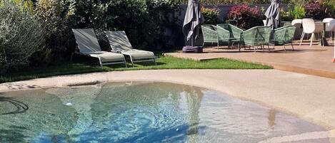 A clear swimming pool with lounge chairs and umbrellas, surrounded by greenery.