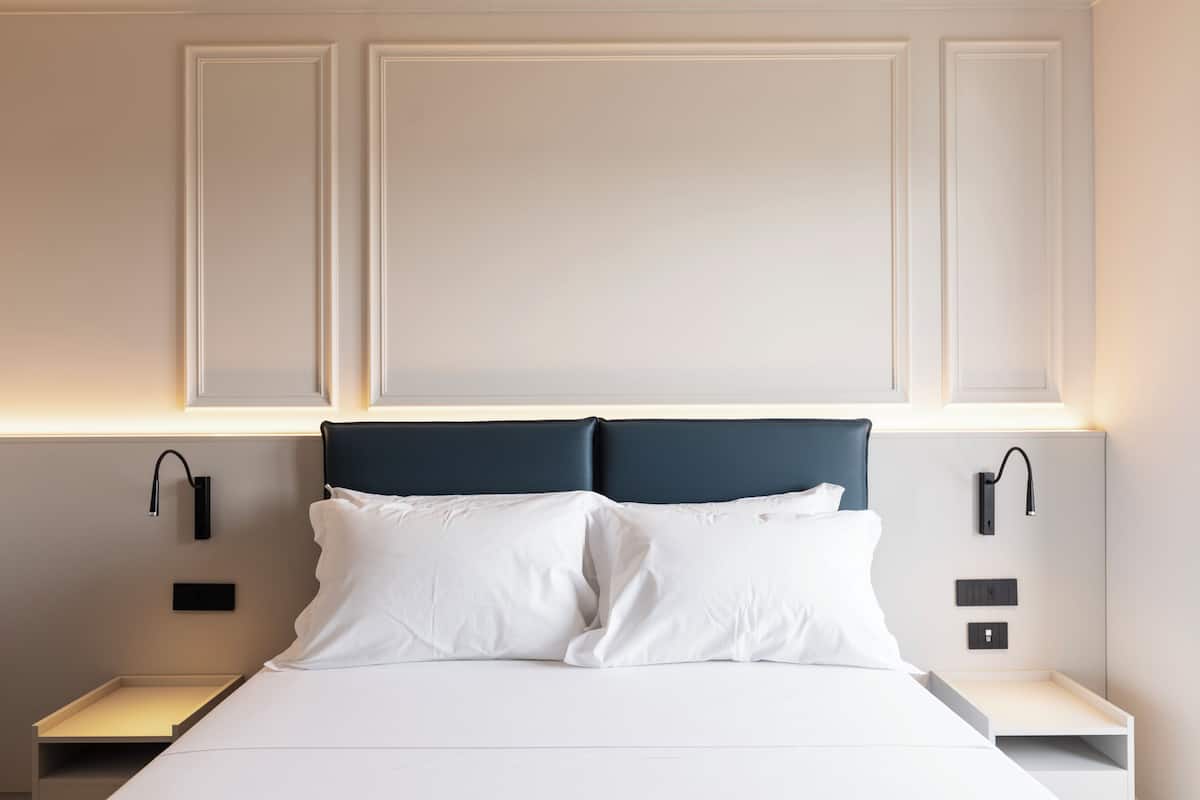Cama bem arrumada com lençóis brancos e cabeceira escura, com mesas de cabeceira laterais com luminárias pretas.
