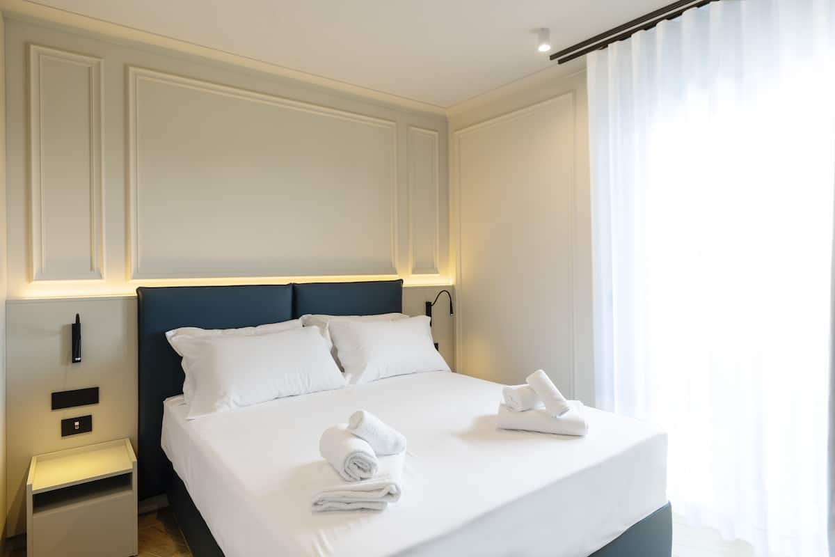 Cama bem arrumada com lençóis e toalhas brancos, cabeceira escura e mesa de cabeceira com abajur.
