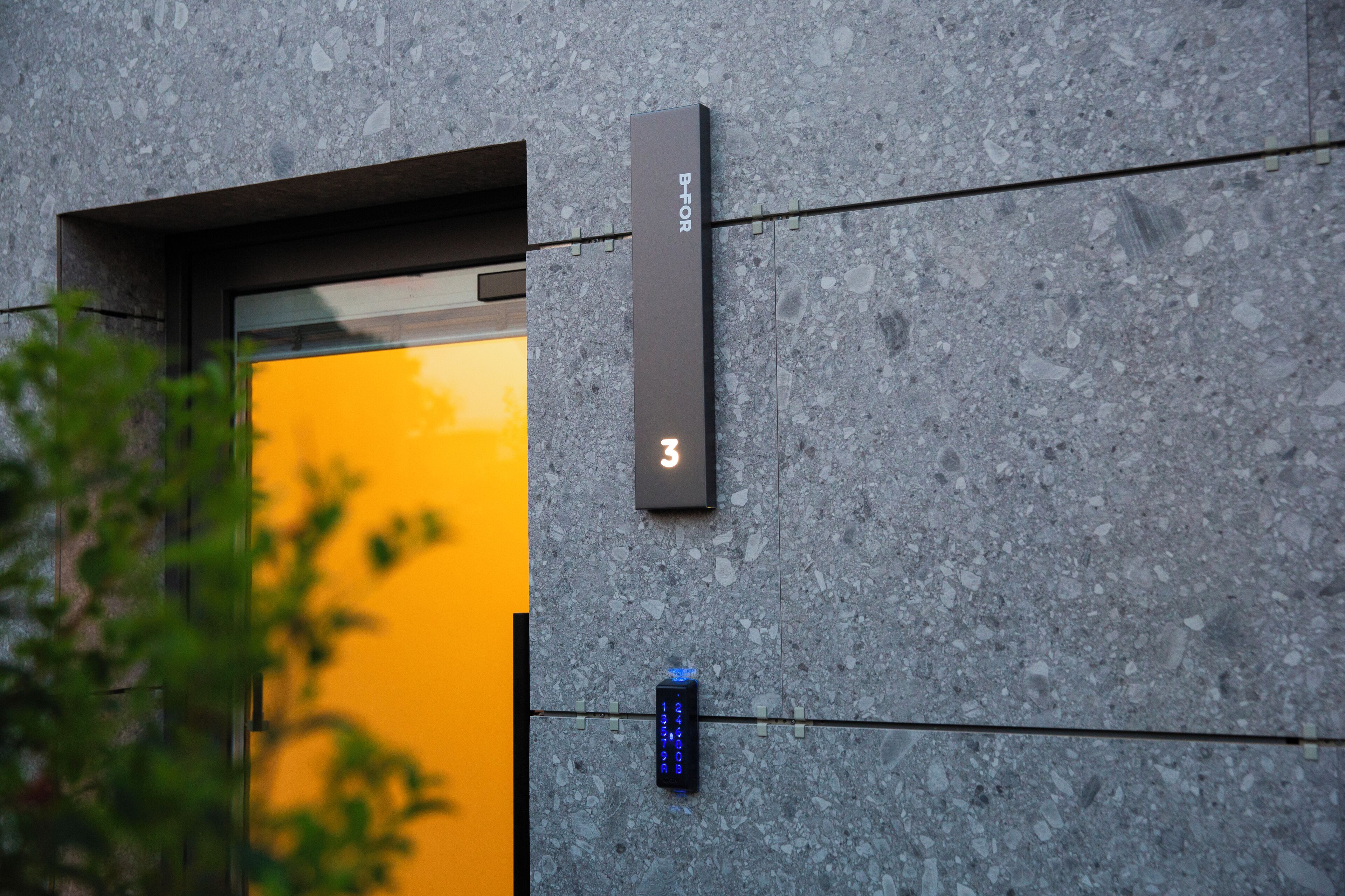 Fachada moderna de un edificio con revestimiento de piedra texturizada, un letrero vertical con las letras 'B-FOR' y un teclado digital para acceso.