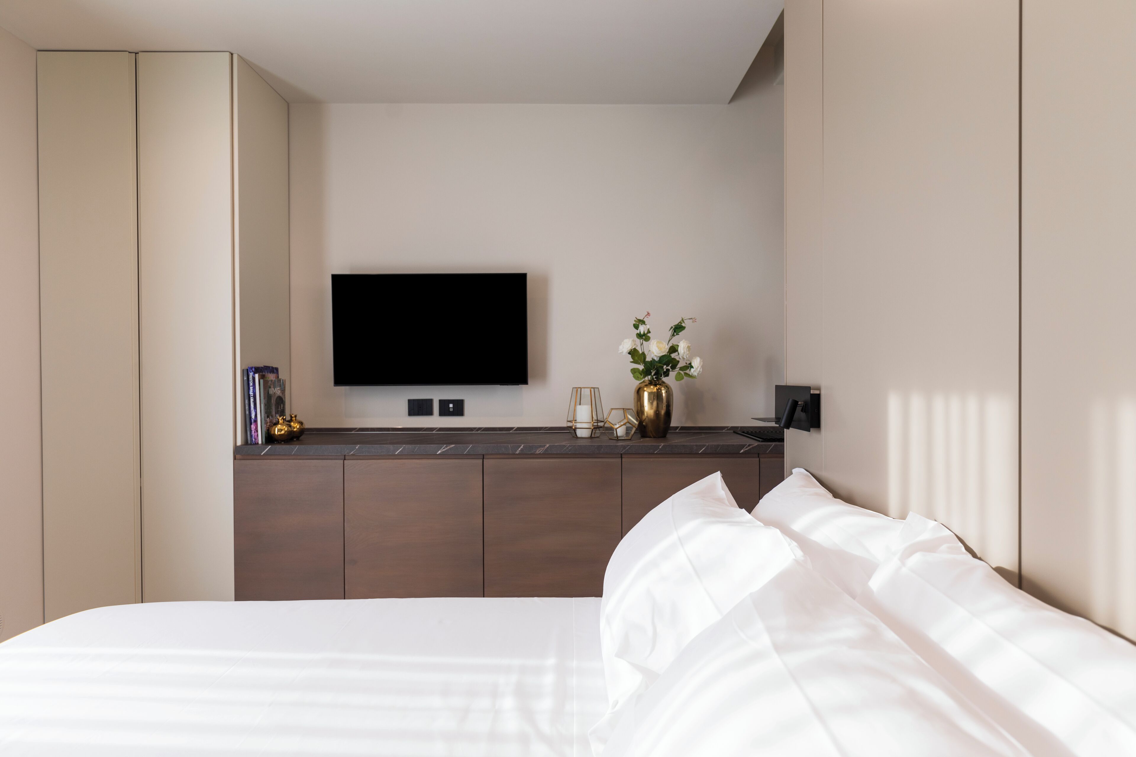 Una habitación de hotel moderna con una cama, un televisor montado en la pared, una mesita con un florero y un armario.