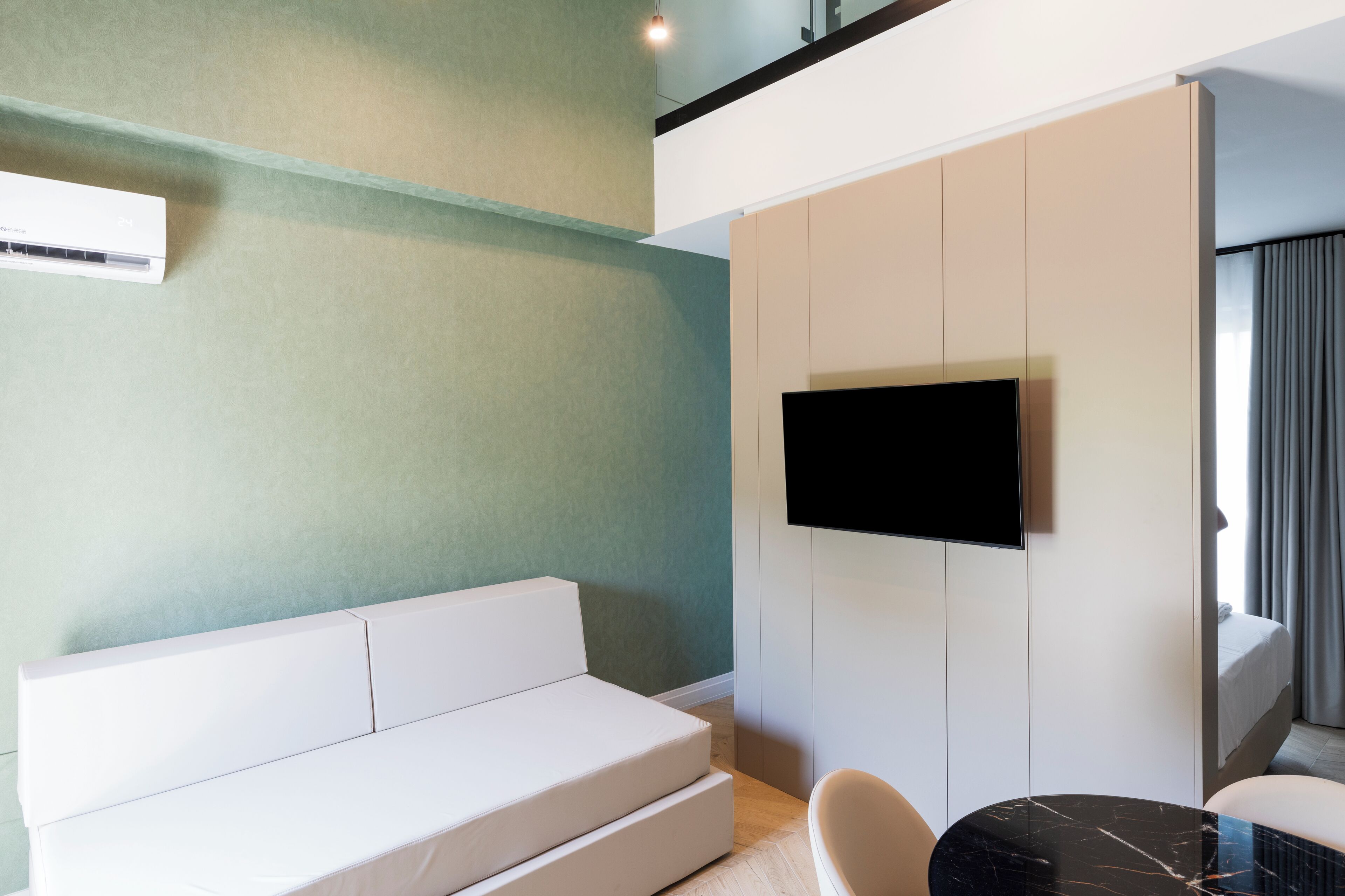 A modern hotel room with a white sofa, a flat-screen TV mounted on a white wall, a small round table, and a window with curtains.
