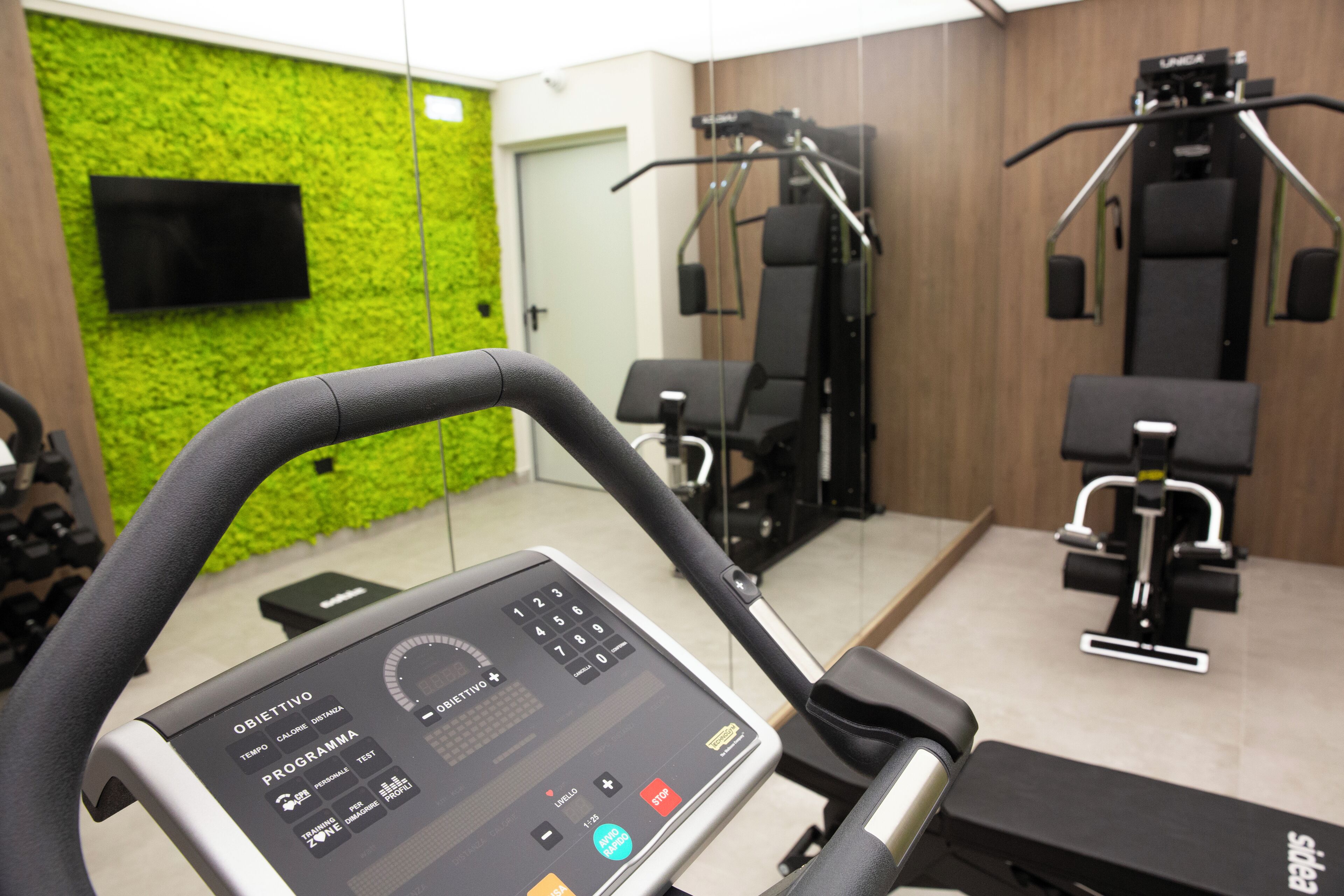 A gym with exercise equipment, a digital interface on the treadmill, and a green wall.