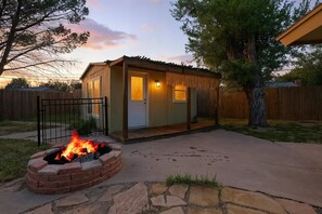 A small wooden cabin with a fire pit in the foreground, a covered porch, and a wooden fence in the background.