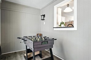Una mesa de fútbolín en una habitación con pared blanca y un espejo que refleja una zona de cocina.