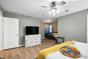 Un dormitorio con cama, televisor sobre un aparador, mesa de comedor con una bandeja de comida y bebidas, ventilador de techo y ventana con cortinas.