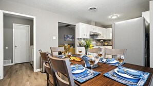 Un comedor con una mesa de madera dispuesta para cuatro personas, sillas y una cocina al fondo.