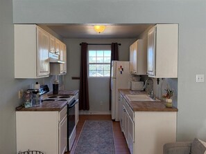 A compact kitchen with white cabinets, a stove, a sink, and a window with curtains.