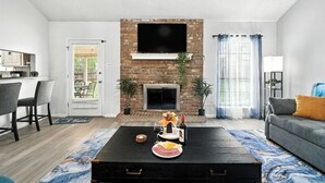 A living room with a fireplace, a television mounted above it, a sofa, and a coffee table with food and drinks. - The Inland Tide 3 bedrooms • 2 bath  • Entire home (Abilene)