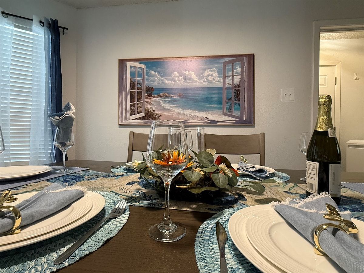 Een eettafel gedekt met borden, bestek en een glas water, met daarboven een schilderij van een strand.