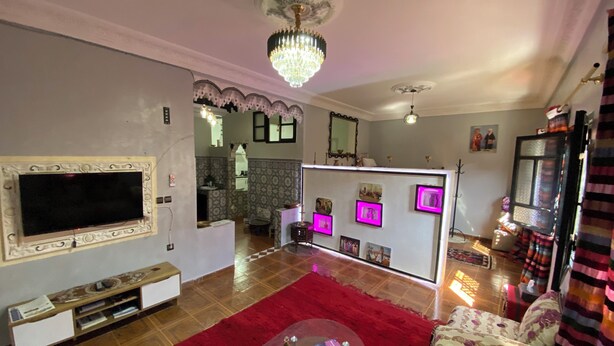 A living room with a TV, a red rug, a sofa, a coffee table, and a chandelier. - Charming Traditional Apartment – Calm & Cozy, Near Marrakech Airport (Marrakesh)