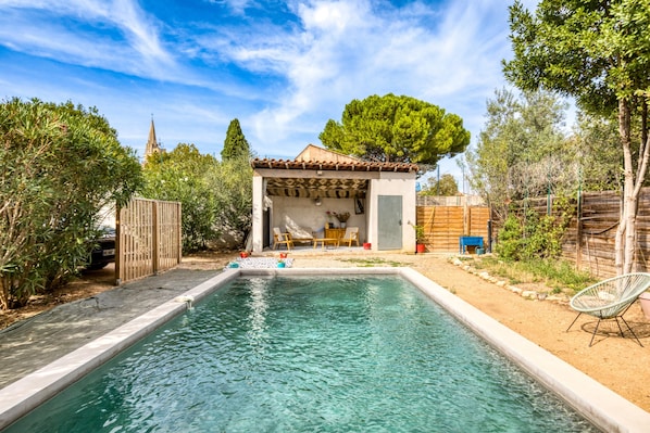 A swimming pool with a covered patio area, a wooden fence, and a view of a church spire in the background.